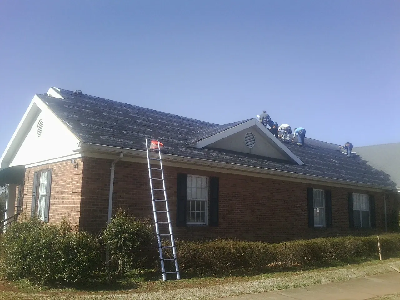 Crew tearing shingles off a brick ranch roof with a ladder set — Metal Roof Repair work by our crew in Flanders