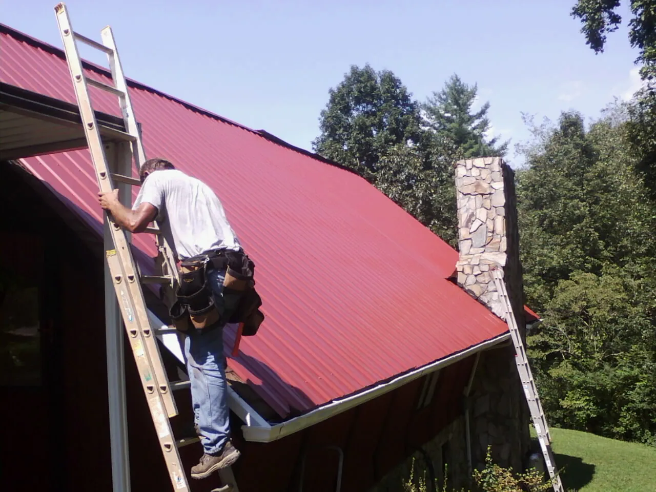 Roofer on a ladder beside a red metal roof and stone chimney — Soffit & Fascia Repair by Roofing Brothers in Flanders
